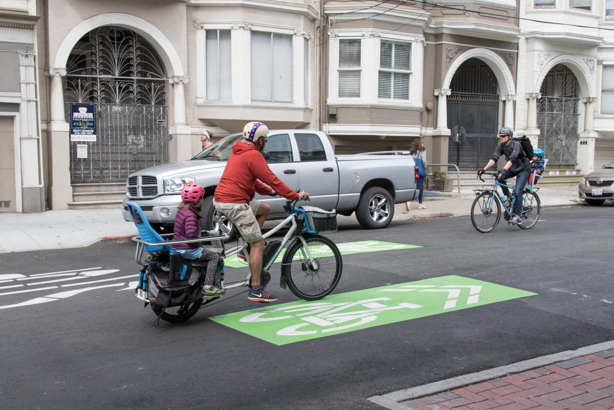 Una persona adulta y un niño andan en una bicicleta para dos personas por una ciclovía pintada de verde que indica que las bicicletas circulan por ahí. 