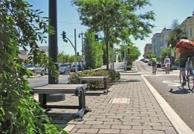 Una persona anda en bicicleta cerca de un área pavimentada con bancas y árboles verdes. 