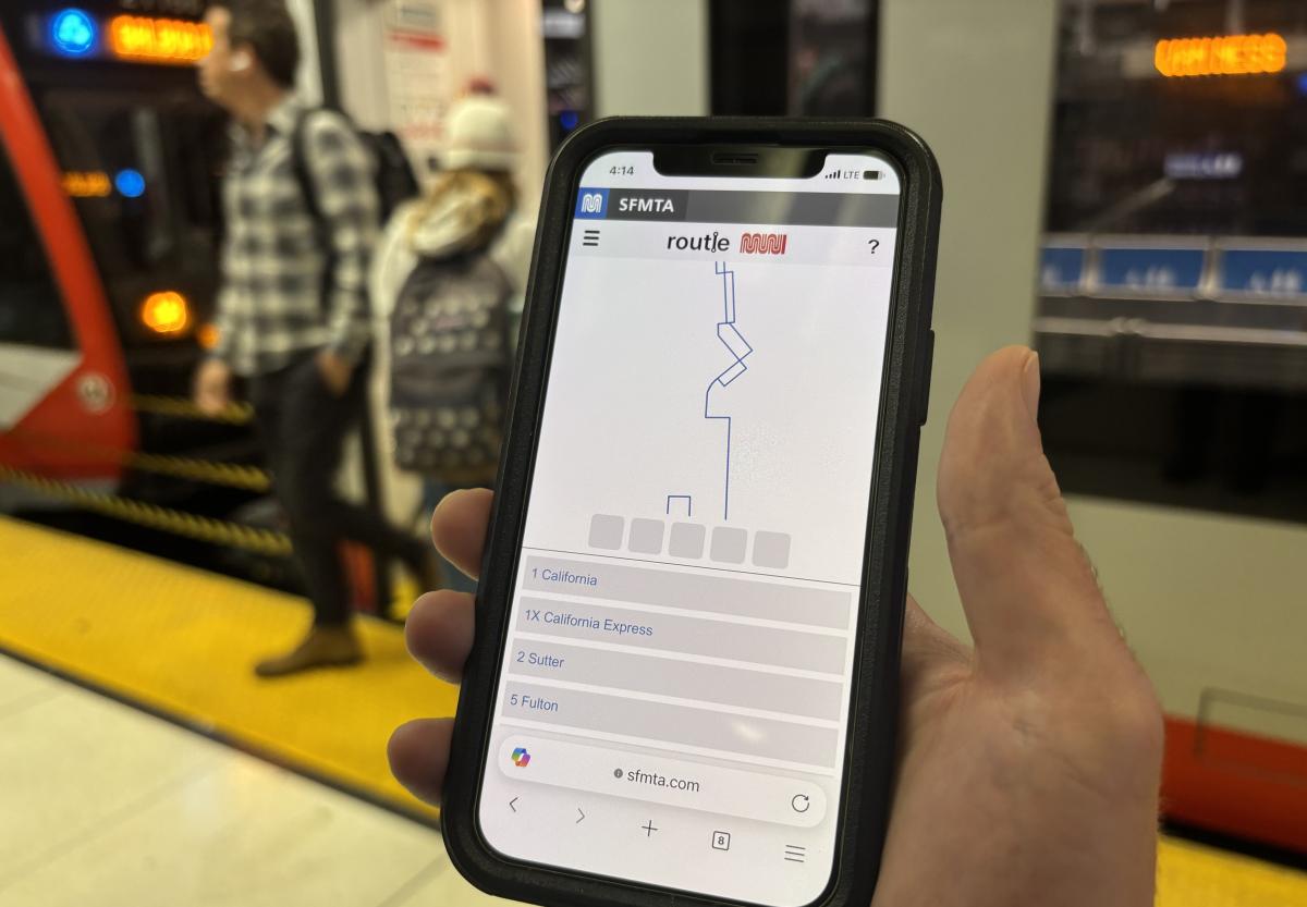 A hand holding a mobile phone in a metro station. A train is in the background.