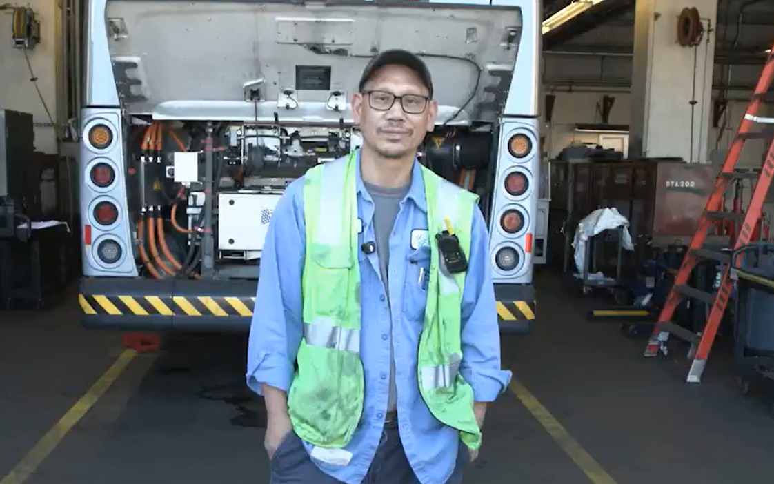 A man wearing a bright vest standing in front of a bus and near a ladder. 
