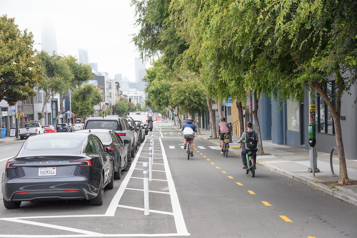 3 Cyclists riding towards downtown in the 2-way parking protected bikeway on Folsom Street. Cars parked in floating parking lane between bikeway and vehicle travel lanes on Folsom Street. Leafy green trees in the sidewalk line the corridor on both sides.