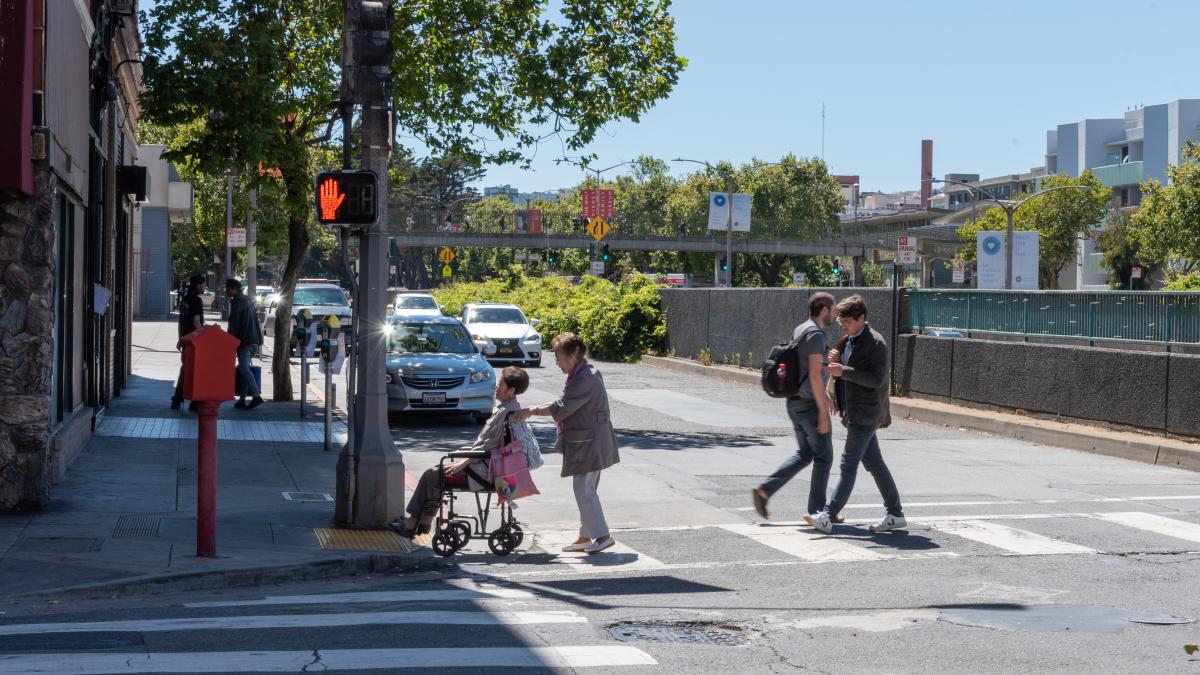 People walking and a senior in a mobility device cross Geary Boulevard in a crosswalk.