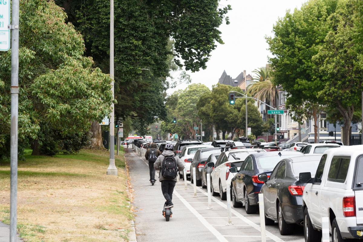 People riding scooters in a bikeway protected from traffic by parked cars on one side. There is grass on the other side of the lane.
