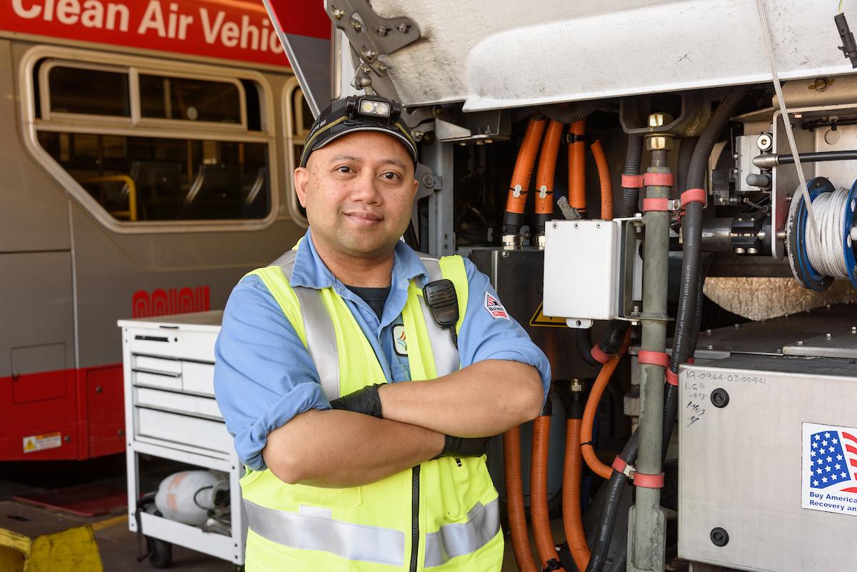 A person wearing a yellow vest and hat while standing near equipment and a transit vehicle.  