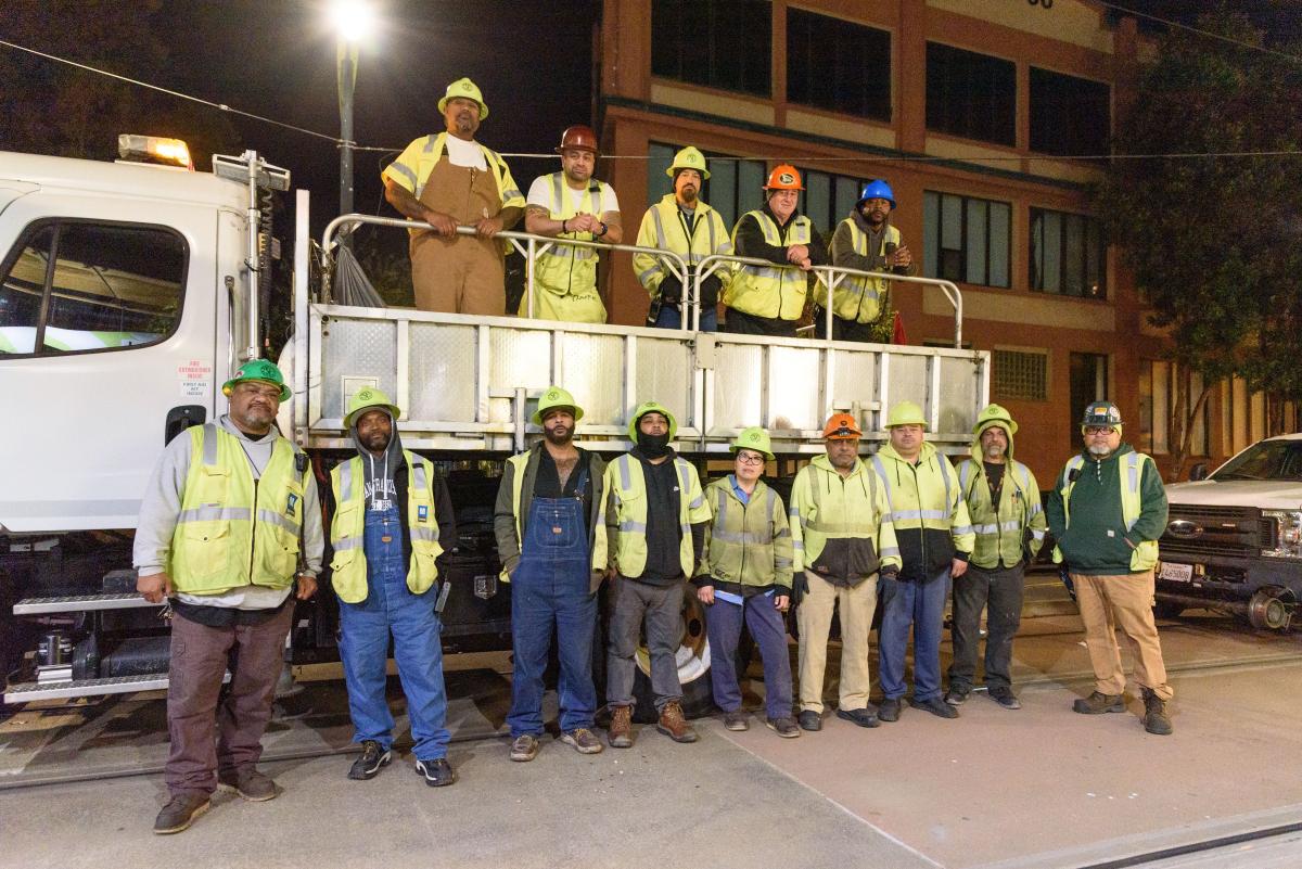 members of Muni's Track Maintenance Team wearing hard hats and standing in a street in front of a truck at night..