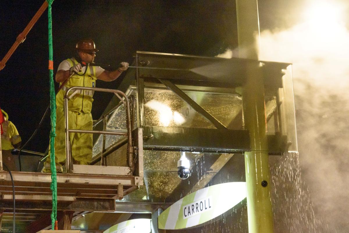 A man wearing a hard hat and standing on a raised platform cleans a transit shelter.
