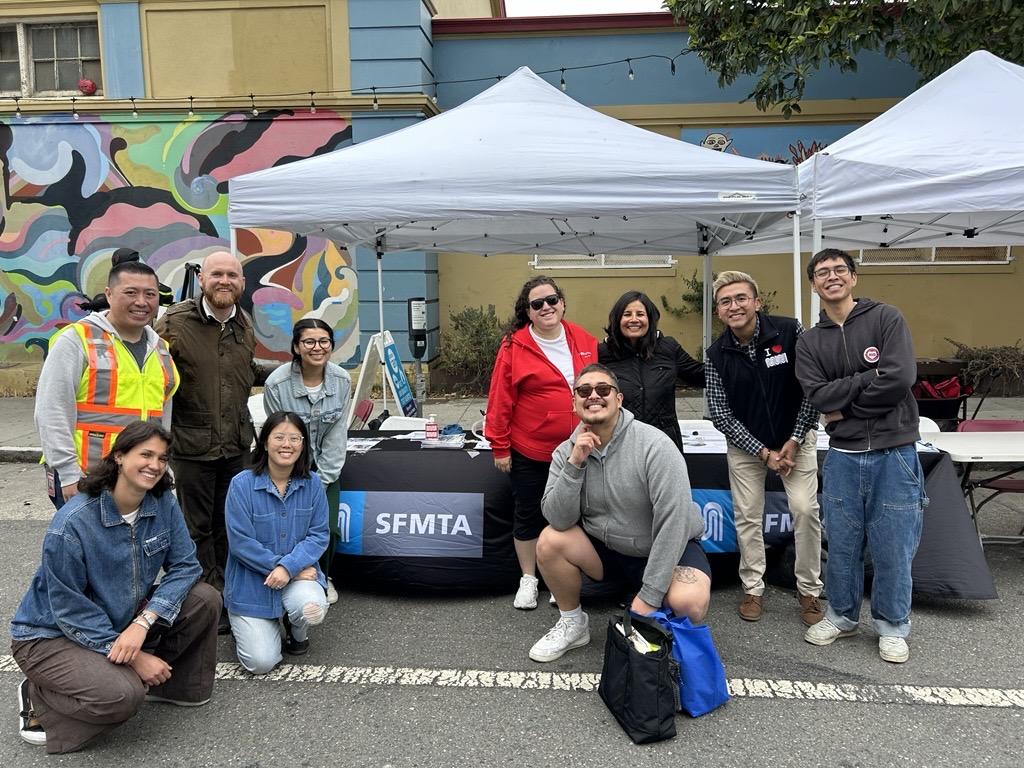 People standing and crouching around a table under a tent. The table has an SFMTA sign draped over it.