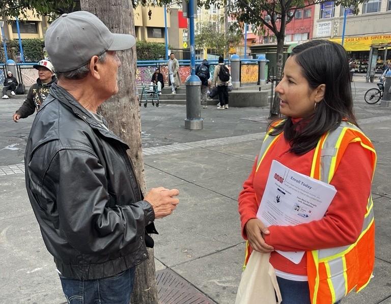Two people talking on a wide sidewalk. One is wearing an orange vest and sweatshirt. The other is wearing a black jacket and baseball cap.