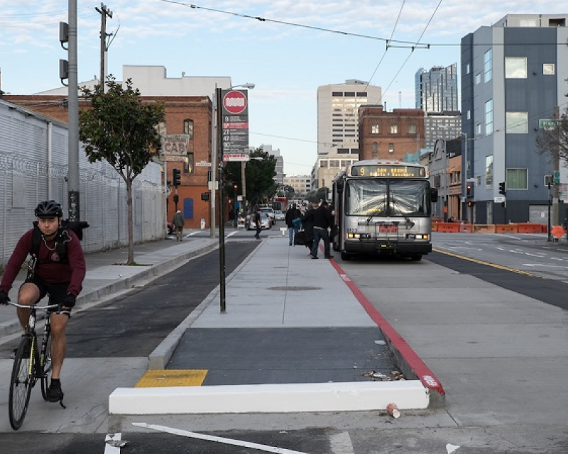 A man rides in a bike lane between the sidewalk and a concrete island where people board a Muni bus on 11th Street at Harrison Street.