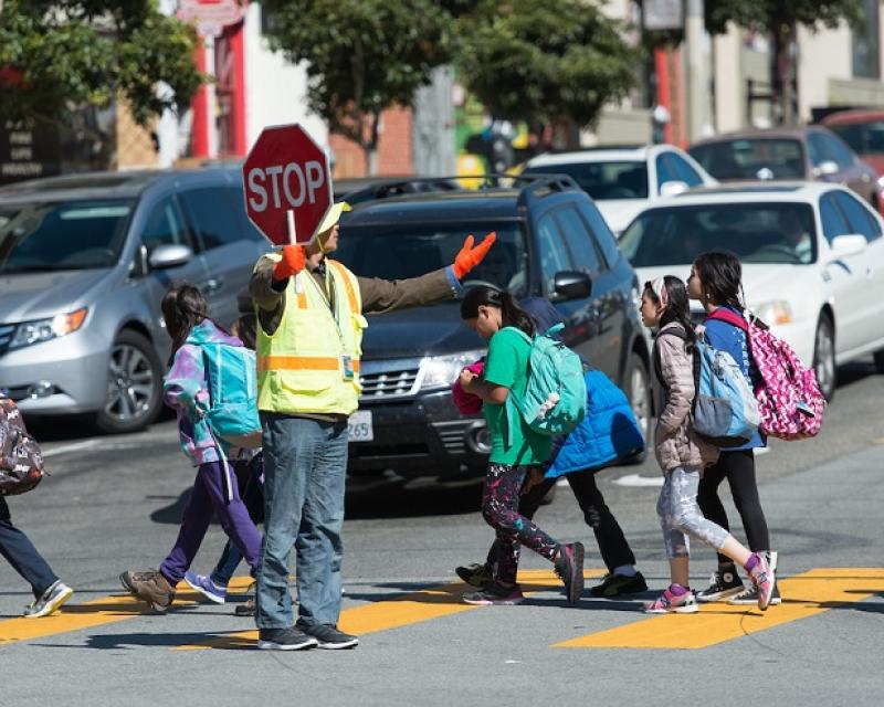 A crossing guard holds a stop sign as children walk in a crosswalk in front of stopped cars.