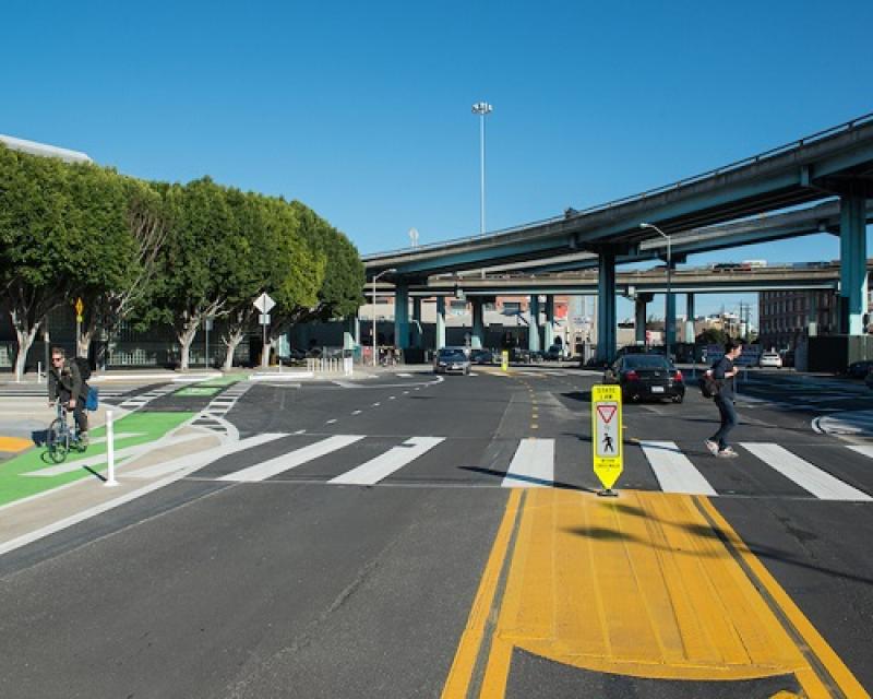 Tree-lined intersection with pedestrians and cars crossing and bright yellow and green markings.