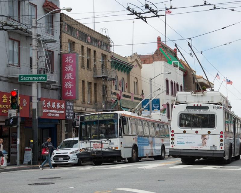 Two Muni coaches passing each other as people use a crosswalk in Chinatown during the day.