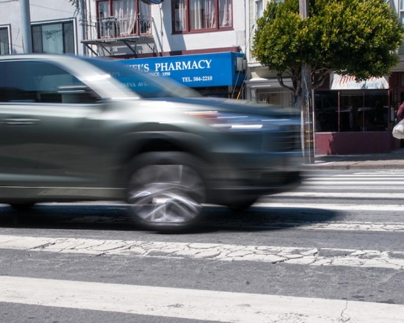 A car appears blurry as it moves through a crosswalk in a San Francisco intersection with pedestrians crossing in the background on Geneva Avenue.