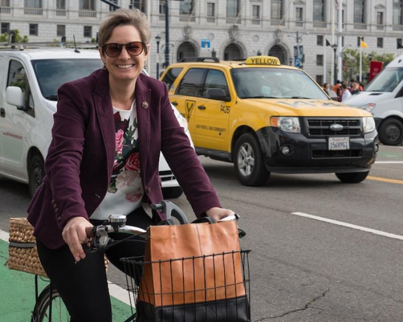 Woman on a bicycle with traffic and Civic Center buildings behind her.