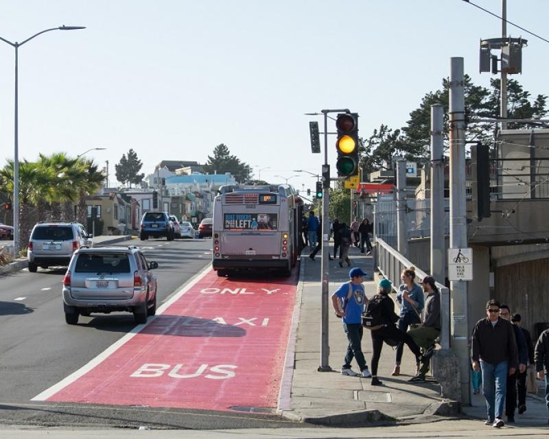 Pedestrians wait to cross San Jose Ave. from Balboa Park Station with Muni bus and cars driving up Geneva Ave. on the left.