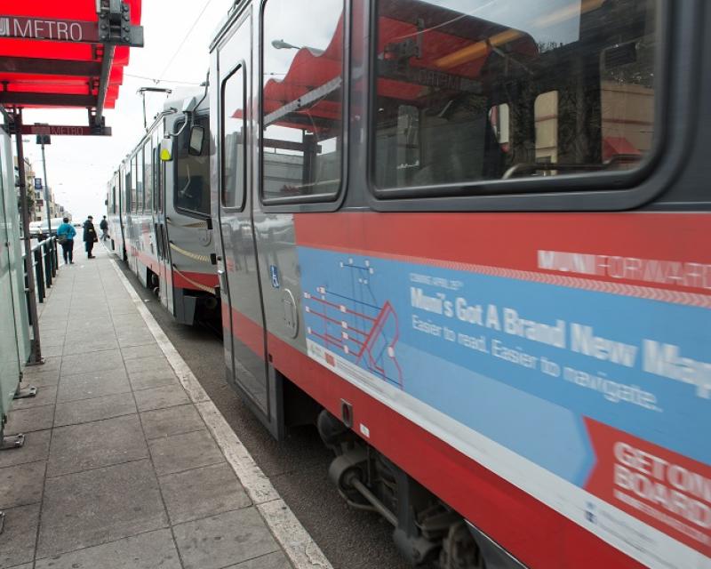 Muni light rail train sits at a transit shelter with a red awning and a Muni Metro logo.
