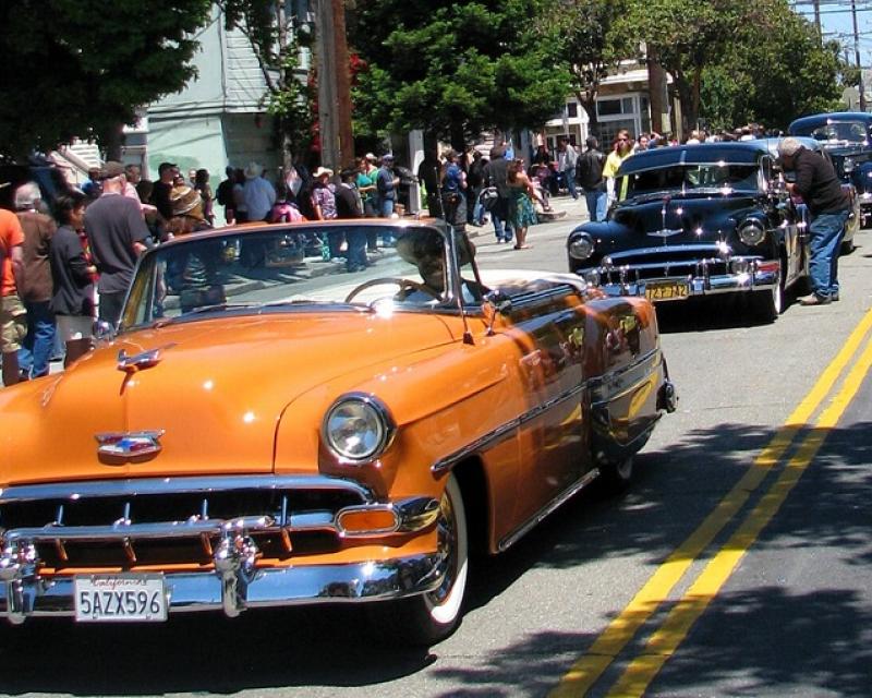 A line of lowrider cars drives down a street with bystanders on the sidewalk in the Mission District on a sunny day.