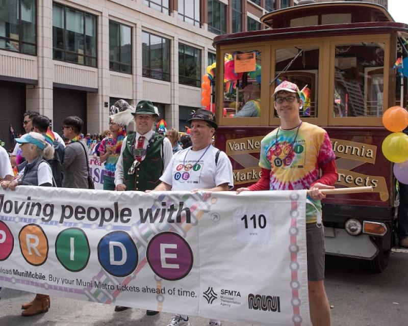 SFMTA staff in the contingent in the Pride parade, standing in front of a decorated motorized Cable Car 62 holding and a banner that says, "Moving People With Pride."