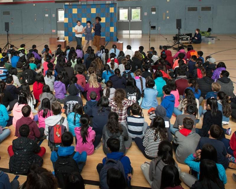 A group of children sit facing three performers on a stage in a gymnasium