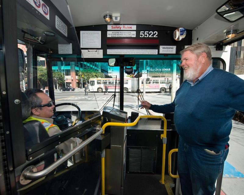 A Muni operator, seated in the bus driver seat, greets a customer with a smile.