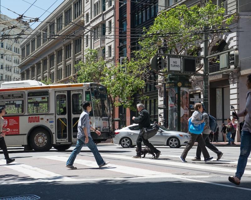 Pedestrians cross a downtown street as a Muni bus and cars cross behind them.