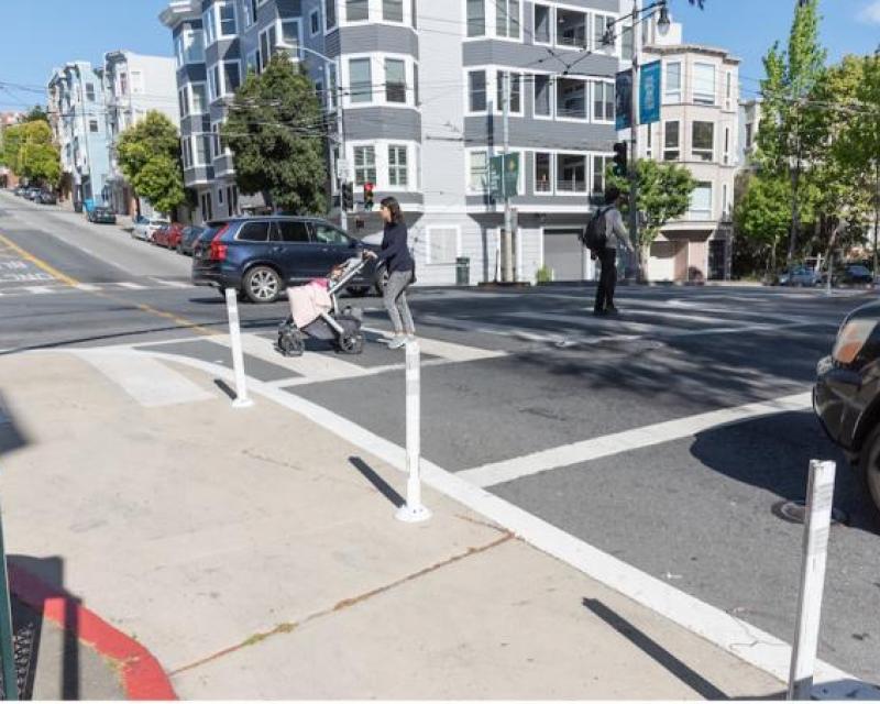 Pedestrians cross in a marked crosswalk toward a painted safety zone next to the curb at the intersection