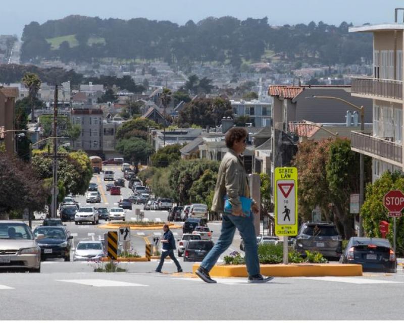 A person crosses a street; behind them there's a concrete pedestrian refuge island down the median.