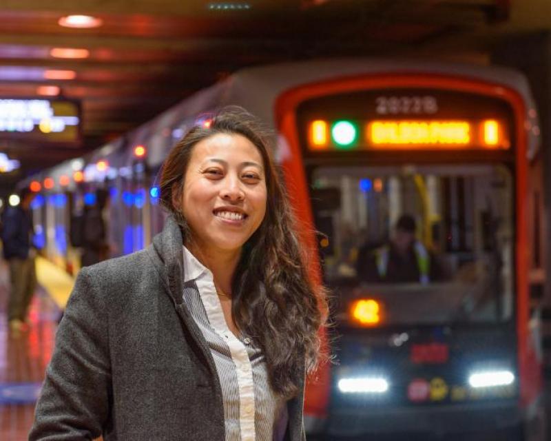 Katherine Kwok smiles on the platform of Castro Station. A train is about to stop in the background.