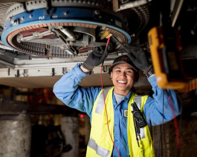 Jeena Villamor holds her hands up to work on overhead equipment for a vehicle. She wears a yellow safety vest and blue uniform.