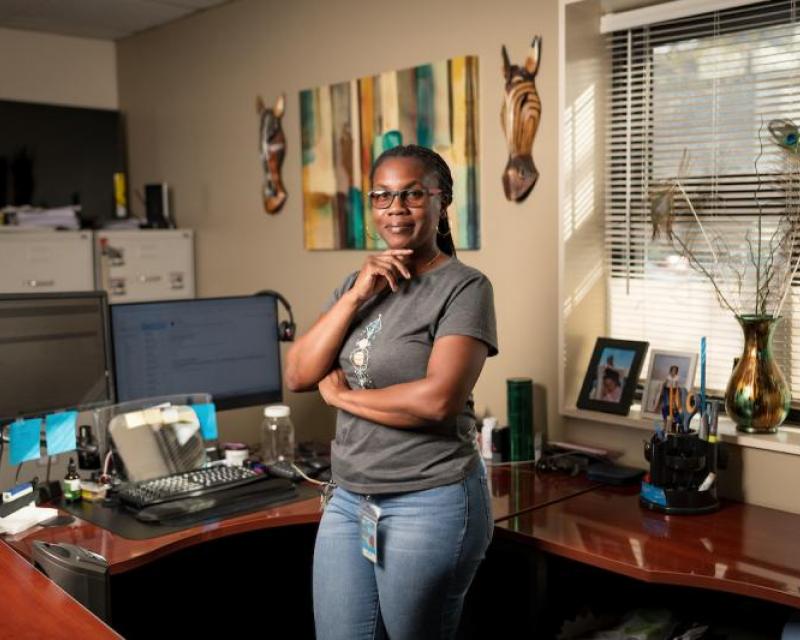 Deneitra Henry smiles from inside her office. We see her desk, computer, photos and other items in the background.