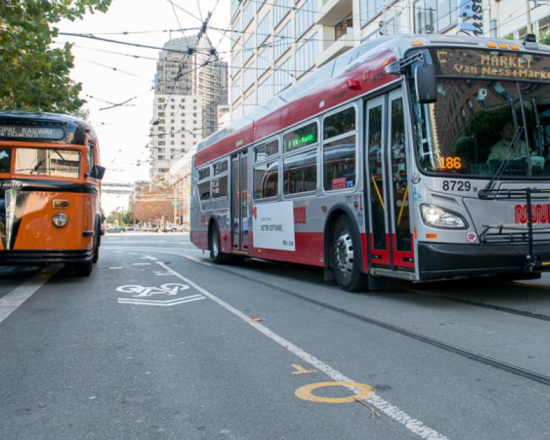 color photo showing two Muni buses next to each other on Steuart street.  On the left is a 1938 orange and black painted White brand bus and on the right is a new 2013 red and grey New Flyer bus.