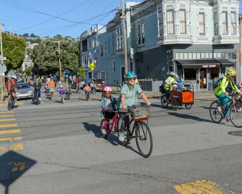 Adults and students ride their bikes, crossing a street on a sunny day.