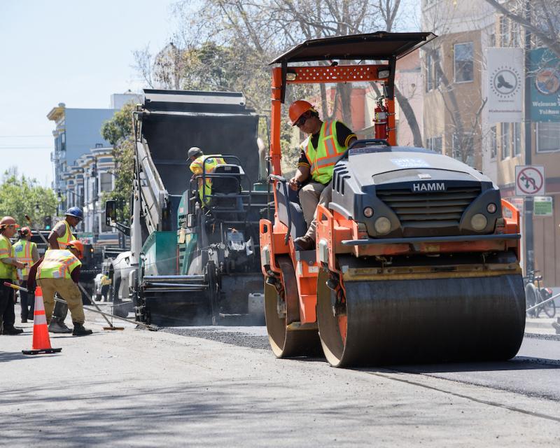 Crews wearing yellow vests repave Valencia Street using large equipment near orange safety cones.
