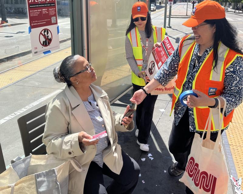 A MuniSafe Day Out volunteer in an orange safety vests speaks to a woman who is sitting down at a Muni bus stop and hands out information to her.