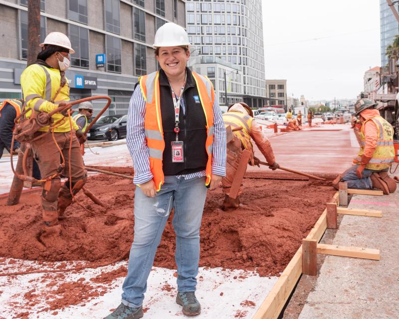 Public Relations Officer Nehama Rogozen smiles for a photo with Van Ness BRT transit lane construction going on in the background behind her.