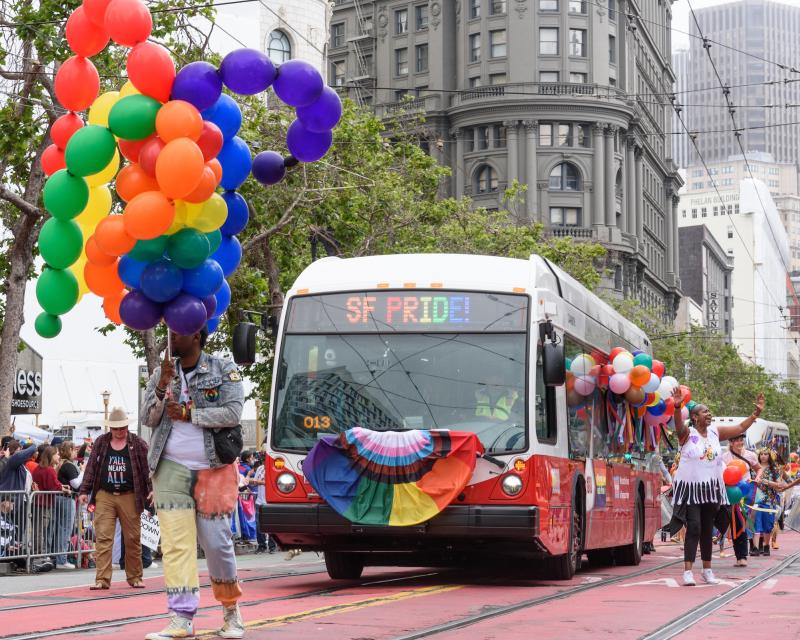 At last year's Pride Parade, a red and white Muni bus that says "SF Pride" and features a rainbow flag drives down Market Street. 