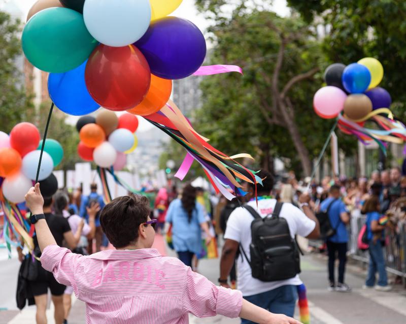 Person holds colorful balloons while walking in the 2024 Pride Parade near other attendees.