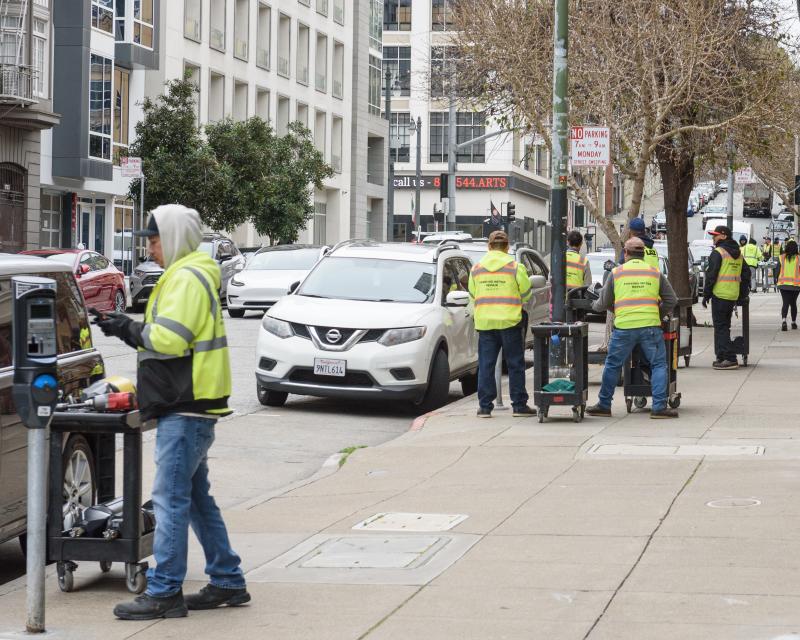 Several crew members in yellow safety vests work to upgrade a few parking meters on a city street.
