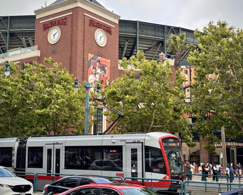 A train in front of a baseball park with people on the sidewalk.