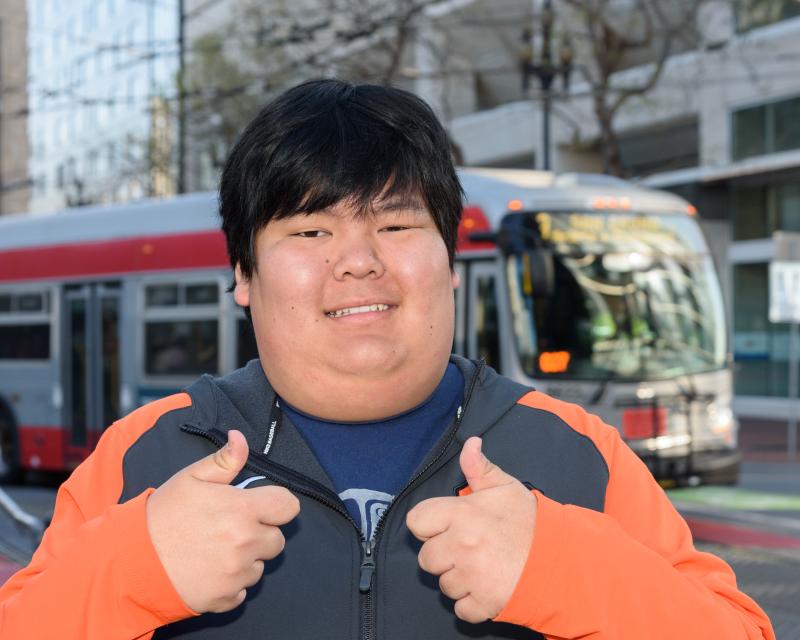 Peter, an Asian young man black hair, is pictured smiling on the street in front of a Muni bus with a “thumbs up” on both hands. He wears an orange and black sweater.