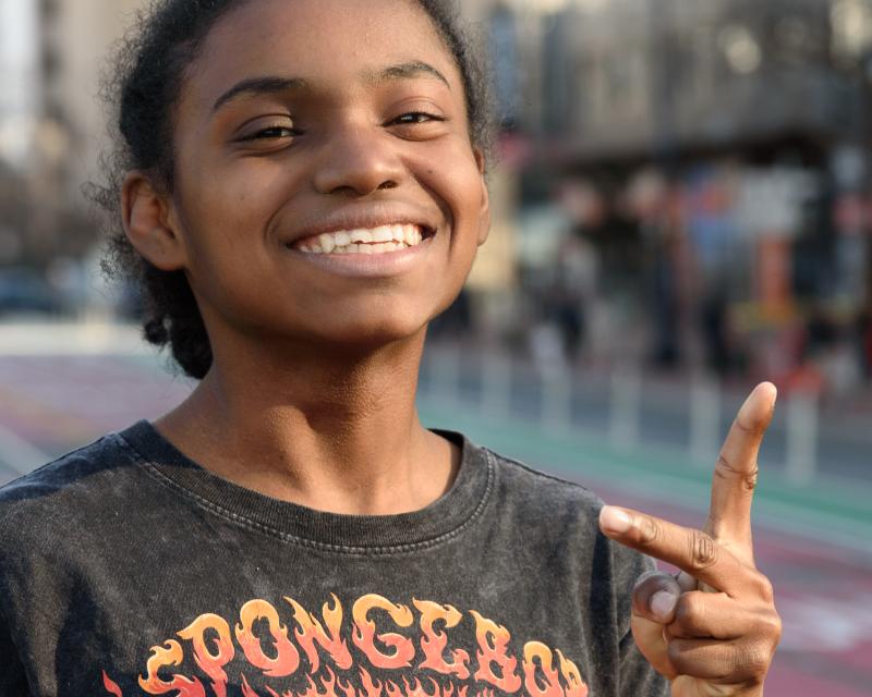Ashanti, a young Black woman is pictured smiling on the street making a peace sign with her hands. She wears a Spongebob tshirt.