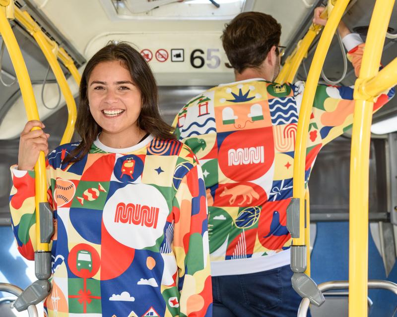 Two people wearing our new Patchwork of the City Holiday Pullover smile on a Muni bus. One faces the camera and the other is turned away, showing the back of the sweater.