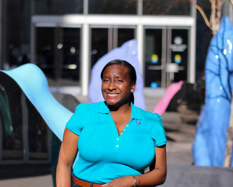 Ammy, a Black woman is pictured smiling in a sculpture garden wearing a bright blue top and jeans. She holds a cane in her righnt hand.
