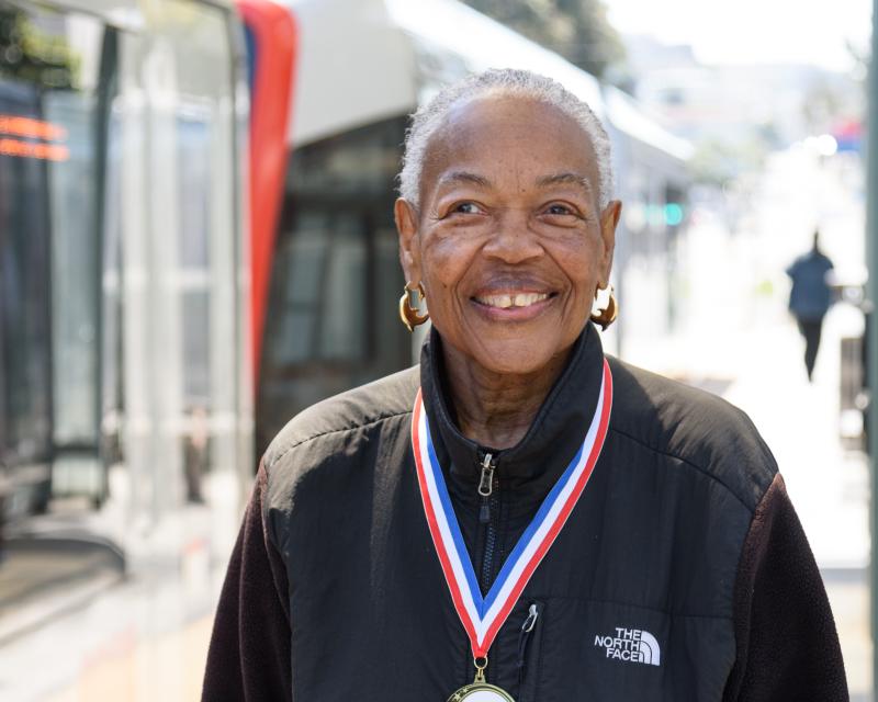 Claire, a gray-haired Black woman is pictured smiling with a medal around her neck. She is wearing a black jacket.  A metro vehicle passes behind her.