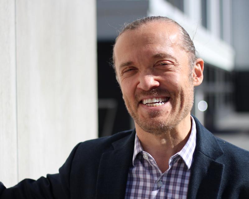 Eli, a White man slicked-back hair, is pictured outdoors smiling and wearing a suit