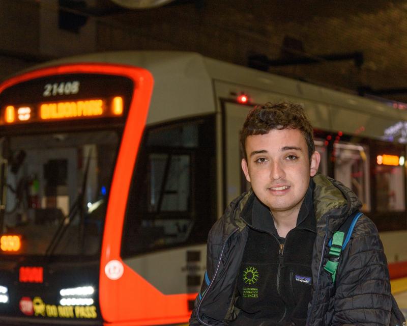 Julian Kanter, a young White man, is pictured with a neutral expression in front of a Muni train as it arrives. He is wearing two black jackets and a backpack