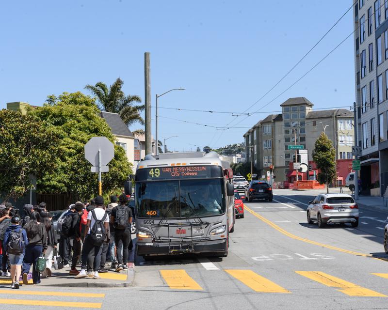 Several young people wait near a corner to board a 49 Van Ness. 