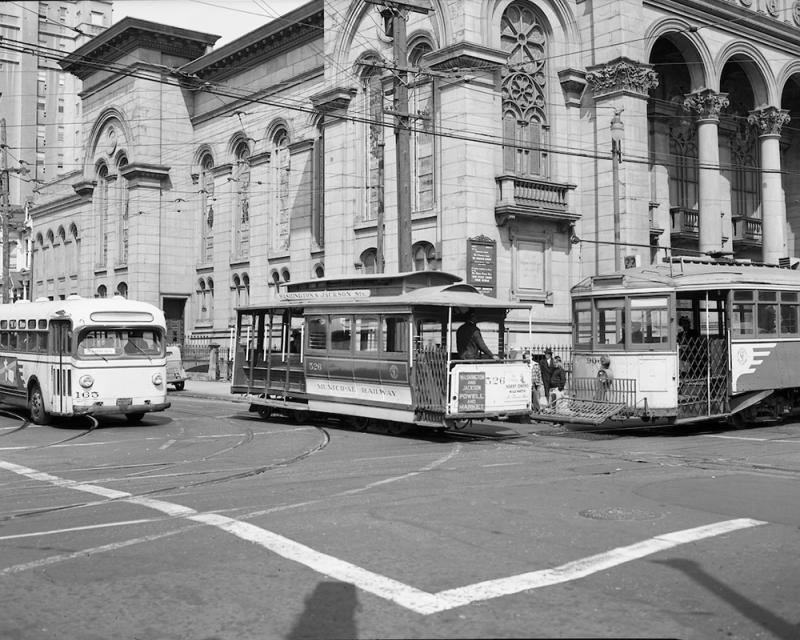 A black and white vintage photo of a cable cars, a vintage streetcar, and a retro bus at an intersection in front of a stone building.