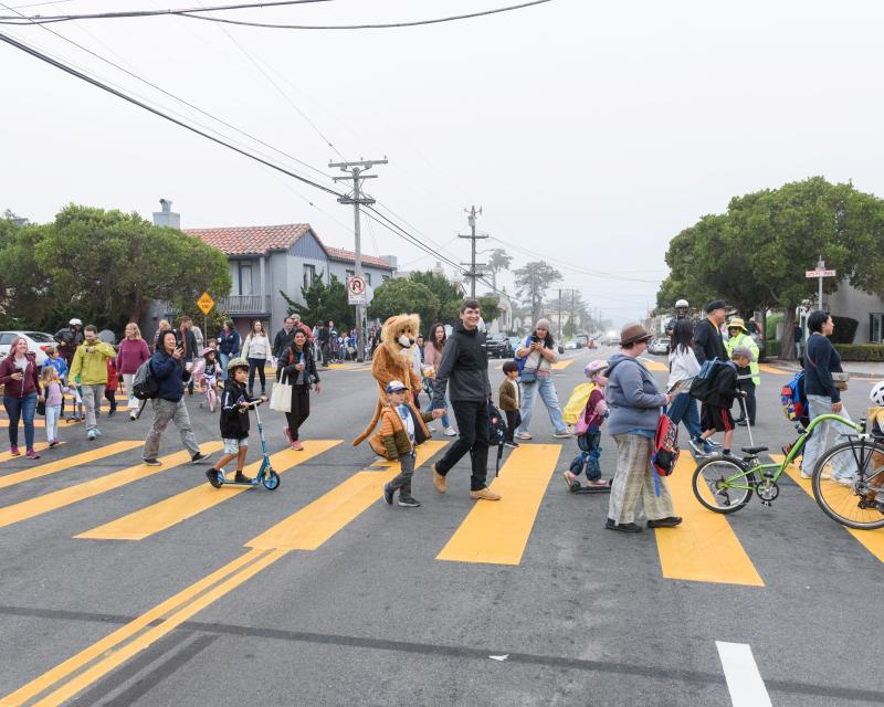 Students, caregivers and others walk and bike in a large group through a yellow crosswalk.