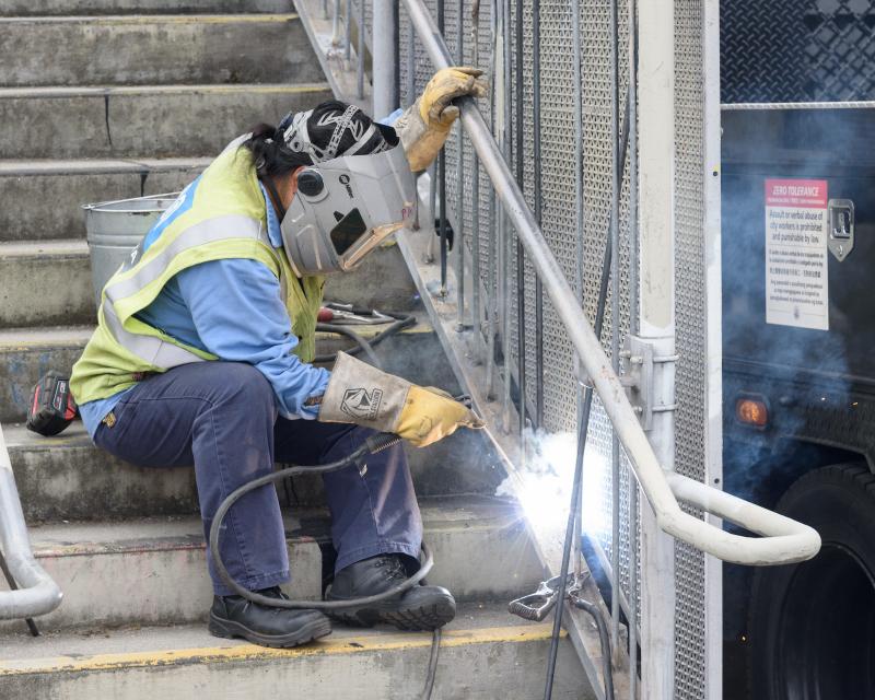 Person wearing yellow safety vest uses a welding tool to repair a staircase railing for our first Fix-It Weekend.